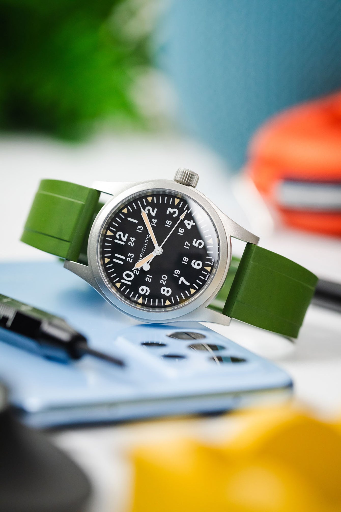 Light reflecting of the glass on a Hamilton Khaki Field watch leant on a blue phone with colourful background on a rubber green watch strap