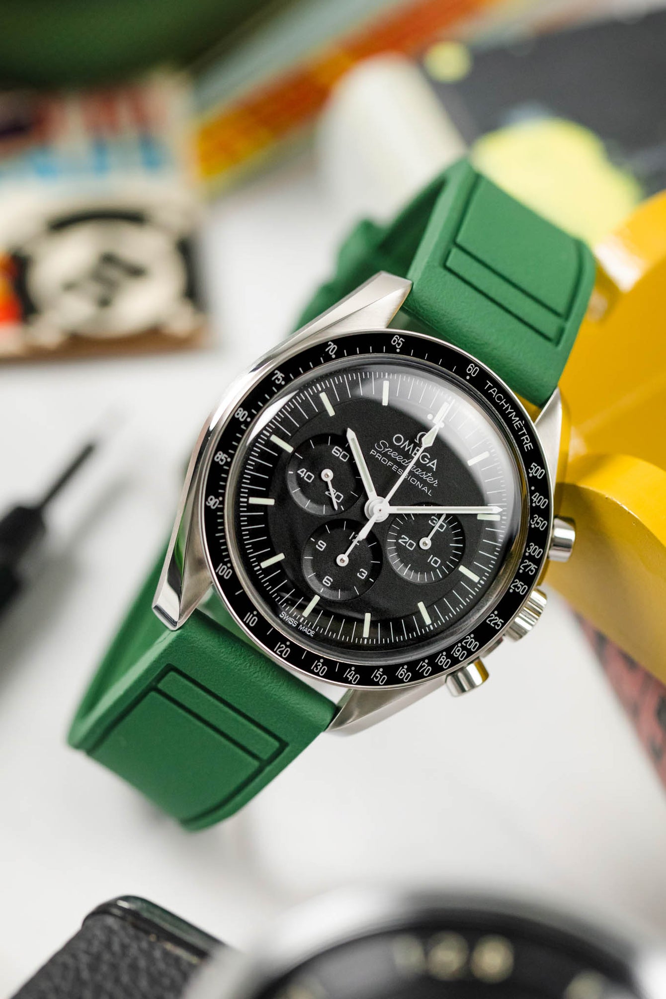 Omega Speedy leant against a yellow wooden trinket box fitted to a green Pilots rubber watch strap