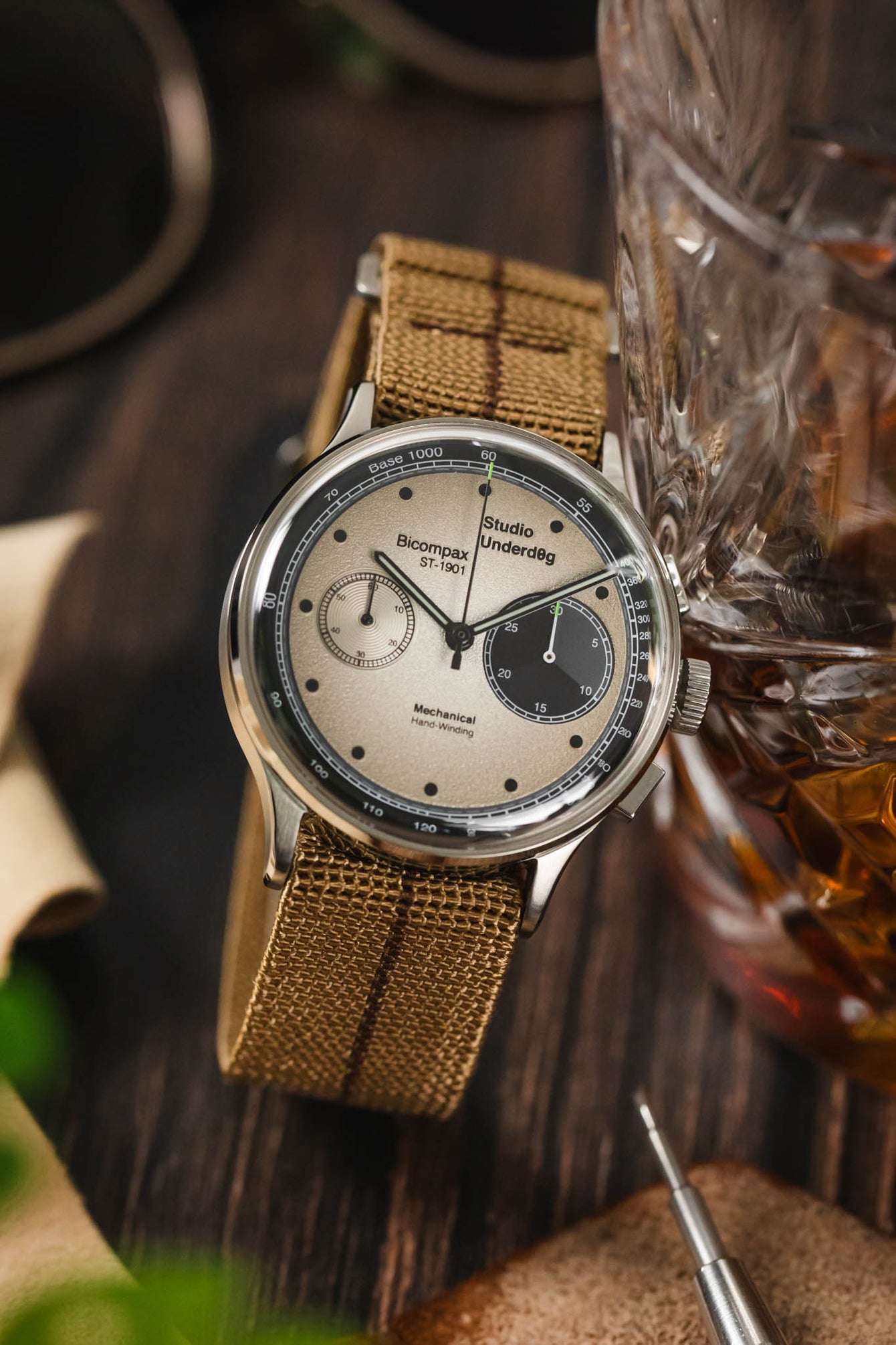 Panda dial watch leant against a whiskey glass fitted to a brown parachute webbing watch strap with brushed stainless steel hardware