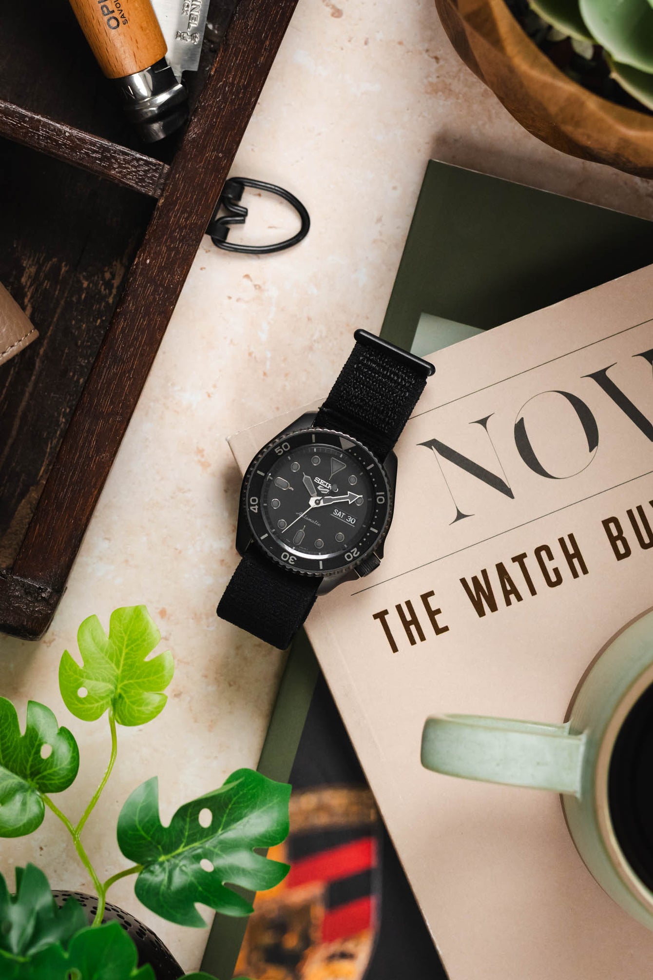 A top-down view of a black Seiko 5 Sports SRPD79K1 watch resting on a magazine, surrounded by a wooden box with a green cup, and artificial plants.